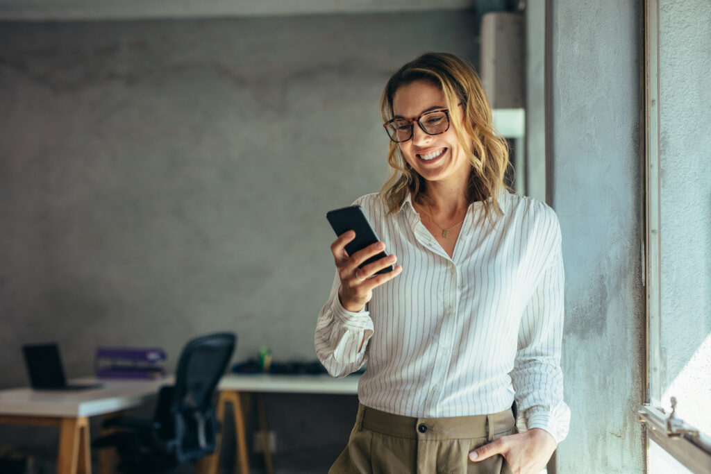 Smiling businesswoman using phone in office. Small business entrepreneur looking at her mobile phone and smiling.